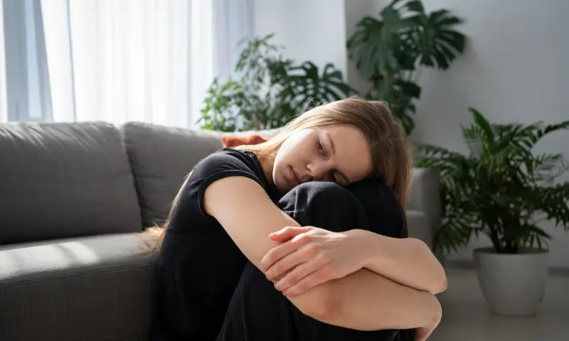 Mulher jovem sentada em um sofá, abraçando os joelhos, com expressão introspectiva, em um ambiente aconchegante com plantas ao fundo, representando momentos de tristeza ou reflexão.