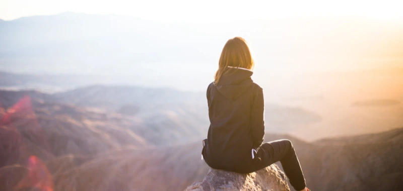 Pessoa sentada em uma pedra, contemplando uma paisagem montanhosa ao pôr do sol, simbolizando introspecção e autoconhecimento.
