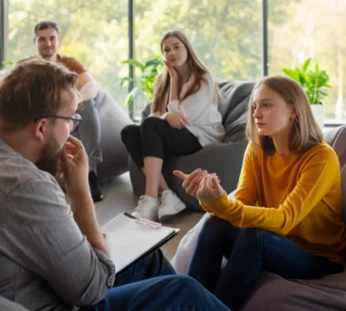 Grupo de pessoas em uma sessão de terapia em grupo, com foco em uma mulher compartilhando suas experiências.